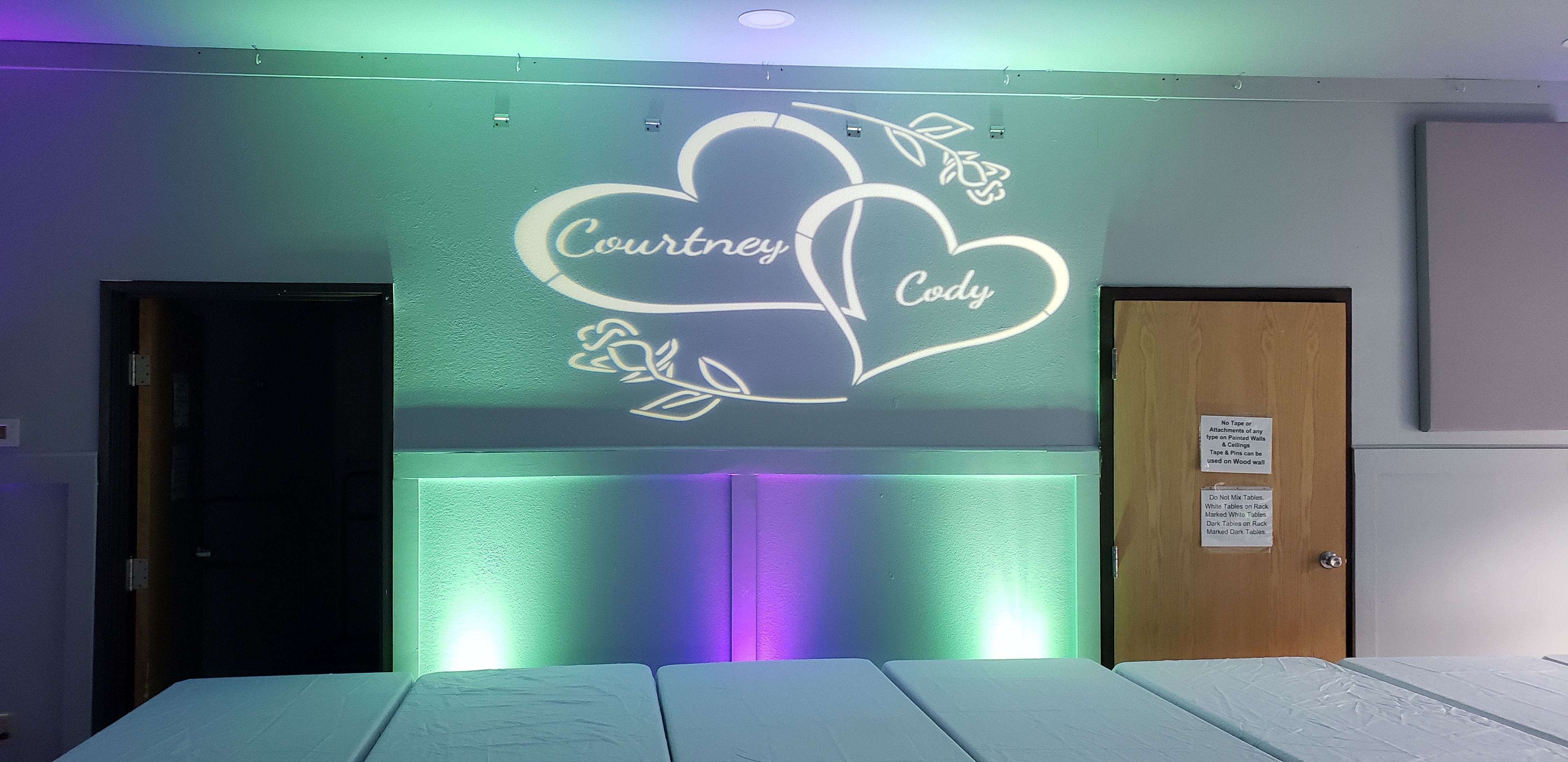 A wedding monogram projected on the wall with purple andmint green up lighting below it.